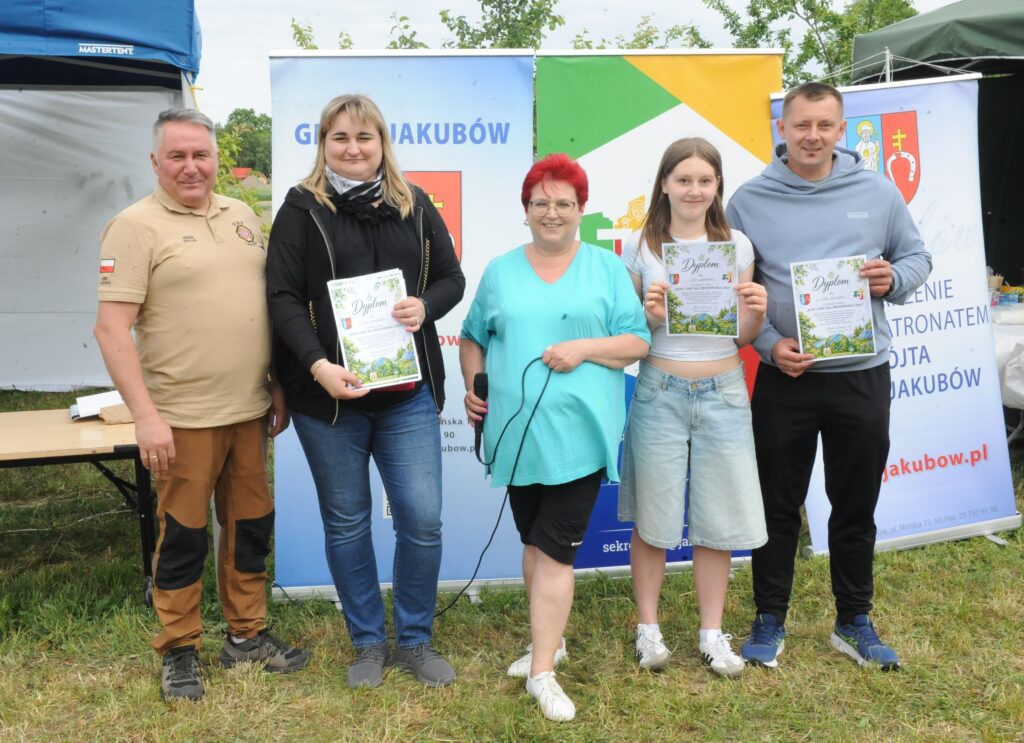 Na zdjęciu jest pięć osób stojących na trawie, za nimi banery. Trzymają dyplomy, a kobieta w środku ma czerwone włosy. Ubrani są w codzienne stroje.