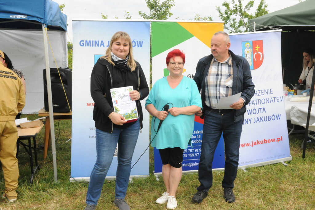 Trzy osoby stoją na trawie przed banerami Gminy Jakubów. Kobieta w czerni trzyma dyplom. Tło jest jasnozielone z namiotem.