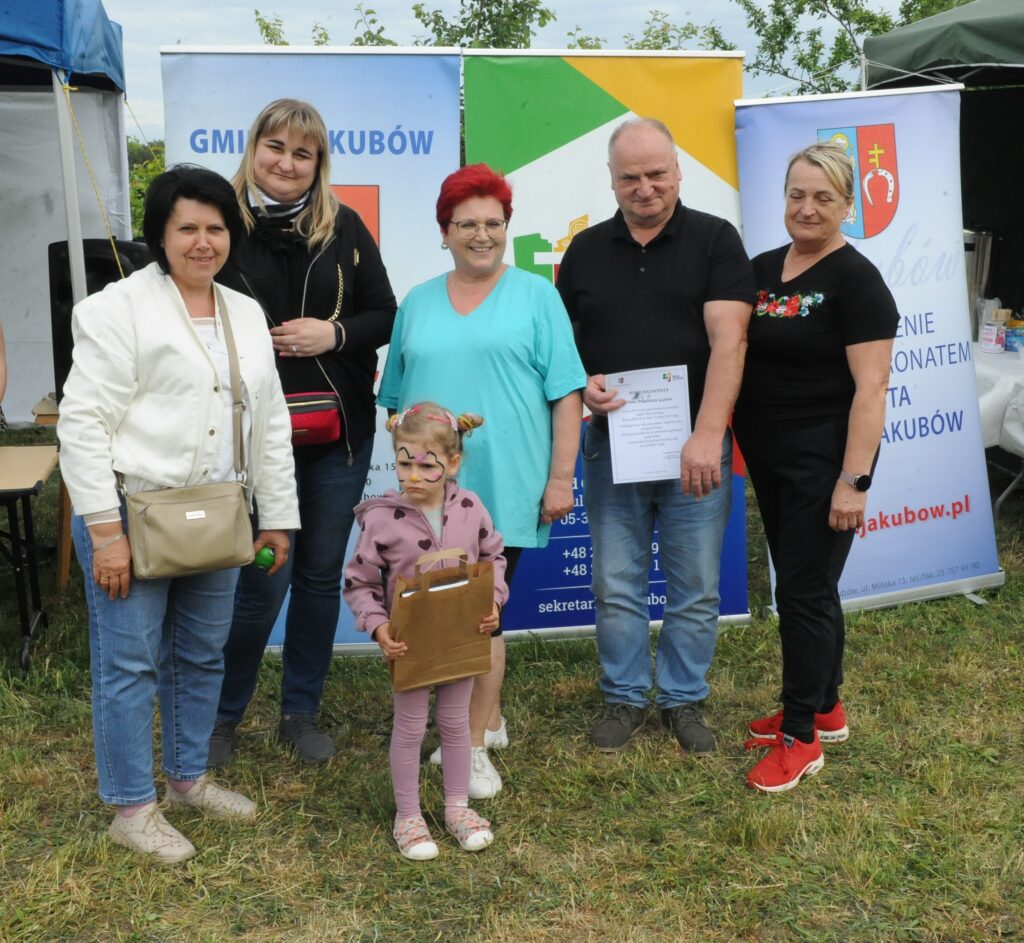 Na zdjęciu znajduje się grupa pięciu dorosłych i jedno dziecko na tle kolorowych banerów. Kobieta w czerwonych włosach stoi w centrum, dziewczynka trzyma torbę. Tło to zielona trawa i jasne niebo.