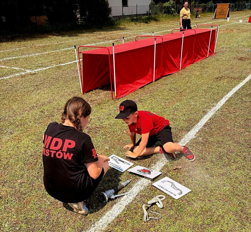 Dwójka dzieci w stroju strażackim układa plansze na trawie, obok czerwone przeszkody. Dziewczynka w czarnej koszulce, chłopiec w czerwonej. Na tle widać boisko z białymi liniami.