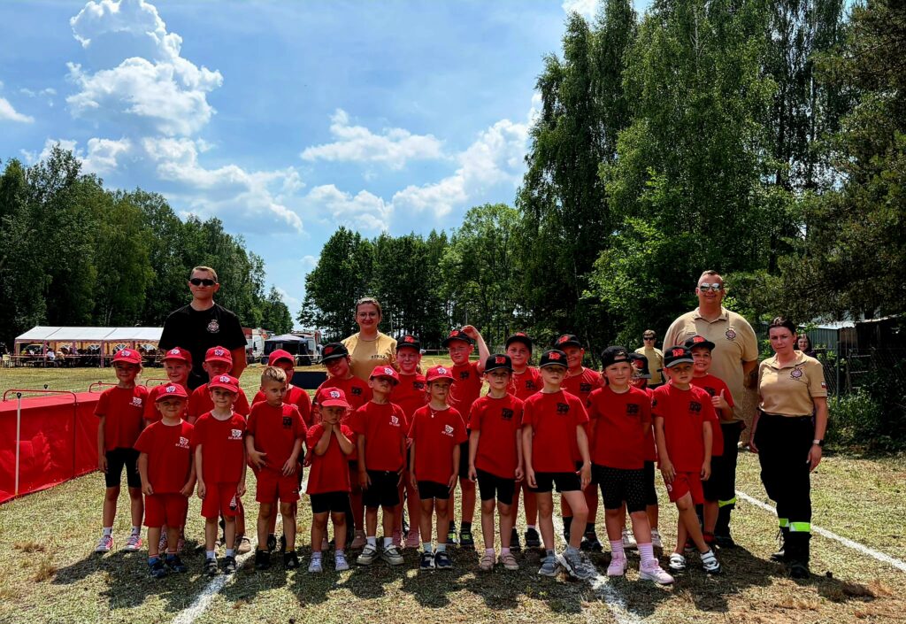 Mała grupa dzieci noszących czerwone koszulki stoi na trawie. W tle są drzewa i błękitne niebo z chmurami. Towarzyszy im kilku dorosłych w beżowych koszulkach.