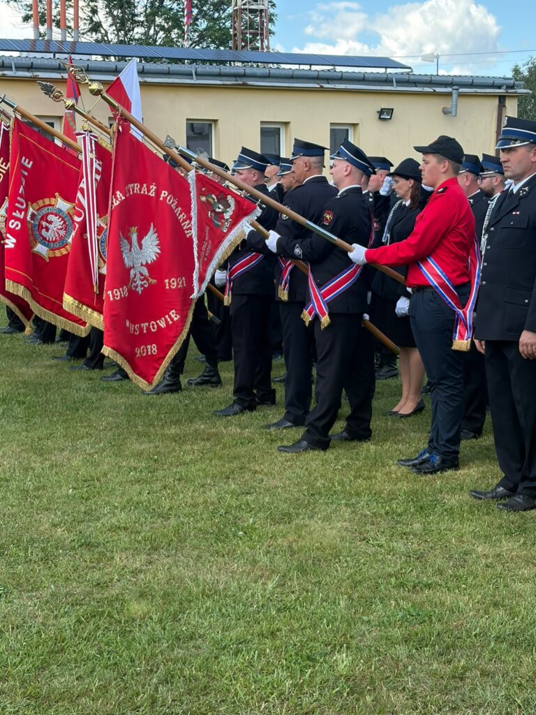 Grupa strażaków w mundurach stoi na trawie z czerwonymi sztandarami. Tło stanowi jasny budynek i niebo z chmurami. Uroczysta atmosfera.