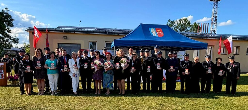 Grupa osób stoi na trawie przed jasnym budynkiem i niebieskim namiotem. W tle powiewają czerwone flagi. Niebo jest błękitne z chmurami.
