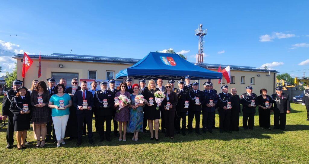 Grupa osób ubranych elegancko stojąca przed budynkiem, w tle niebieski namiot i słoneczna pogoda.