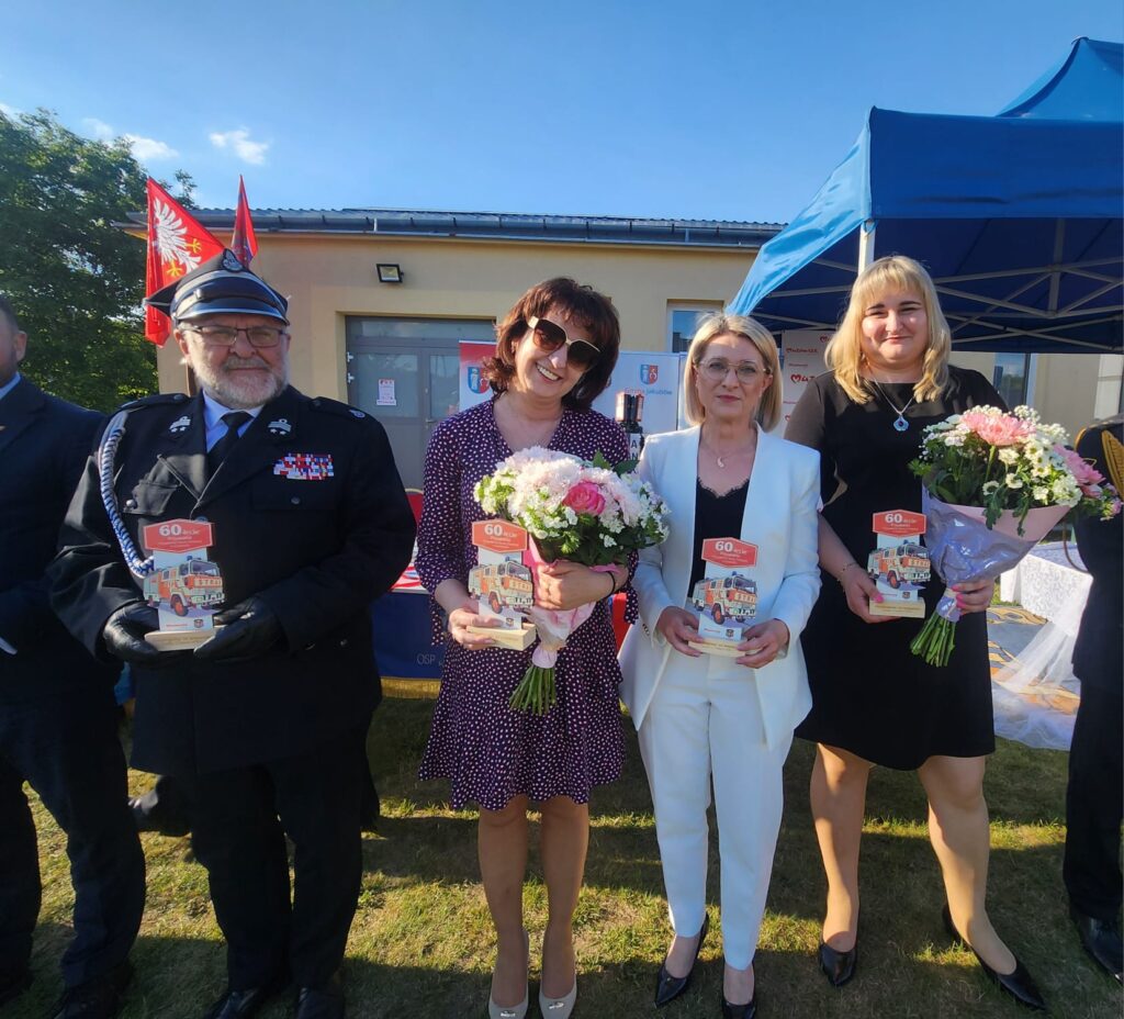 Na zdjęciu widać cztery osoby stojące w plenerze, z kwiatami i dyplomami. Tło to jasny budynek i niebieski namiot. Osoby są uśmiechnięte, a niebo jest bezchmurne.