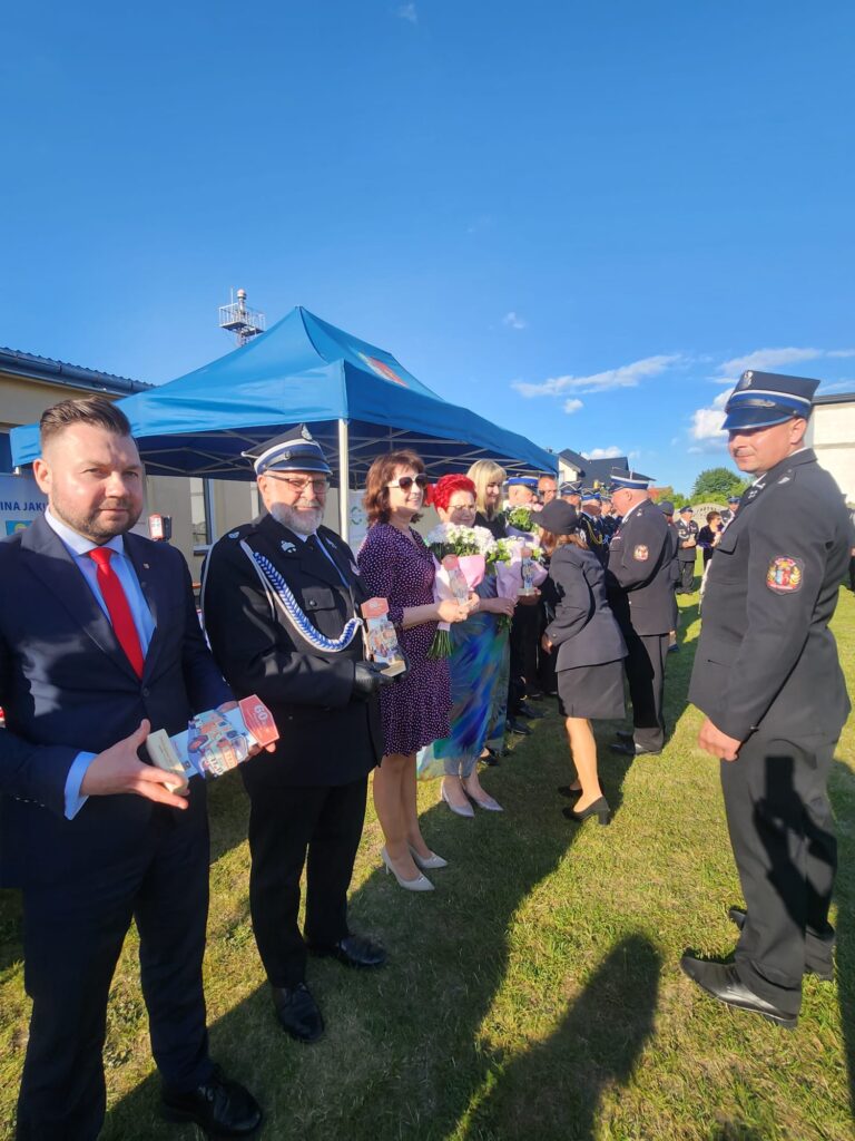 Grupa ludzi w eleganckich ubraniach na trawie, z niebieskim namiotem w tle. Kobiety trzymają kwiaty, mężczyźni w mundurach strażackich. Jasne, słoneczne niebo.