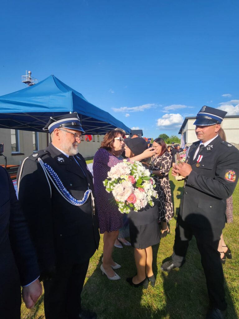 Na zdjęciu są strażacy w mundurach, kobiety z bukietem kwiatów. W tle niebieski namiot i budynek na tle nieba.