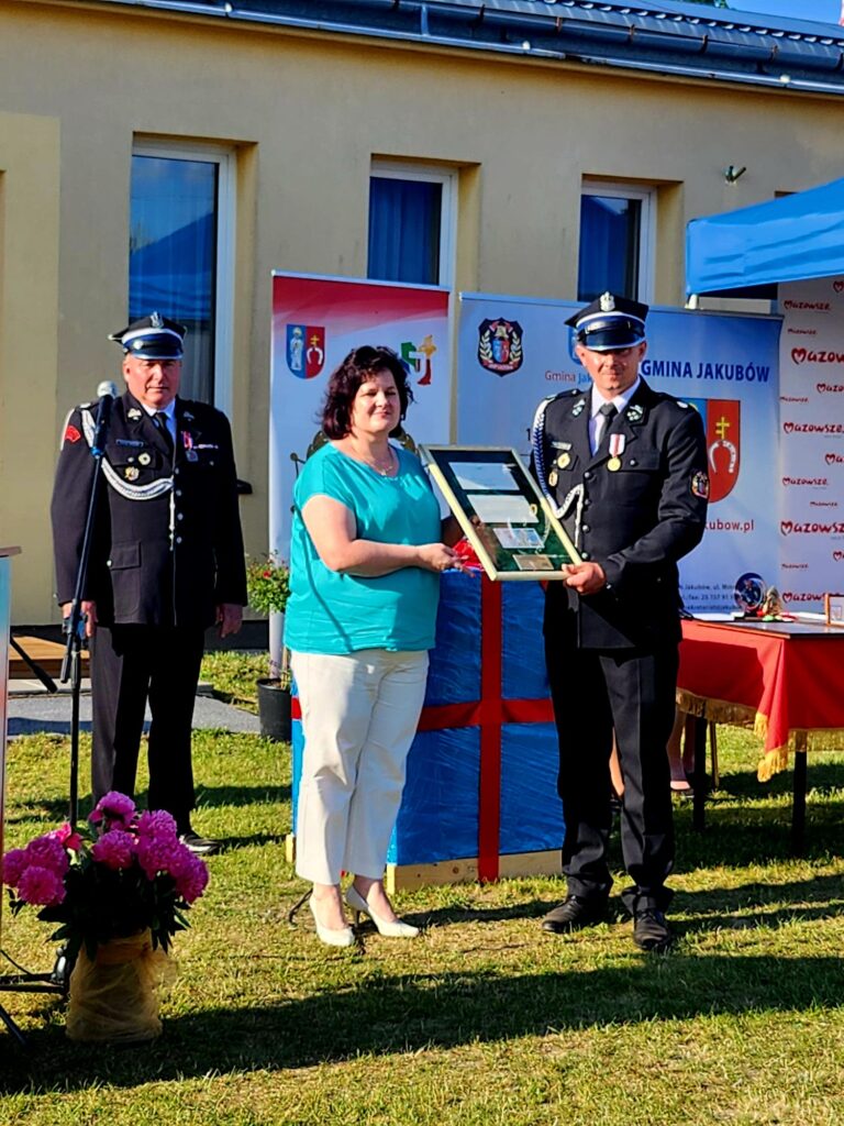 Trzy osoby na zewnątrz, dwie w mundurach, jedna w niebieskiej bluzce, trzymają dyplom. Tło z żółtym budynkiem i banerem. Na trawie stoi kosz z różowymi kwiatami.