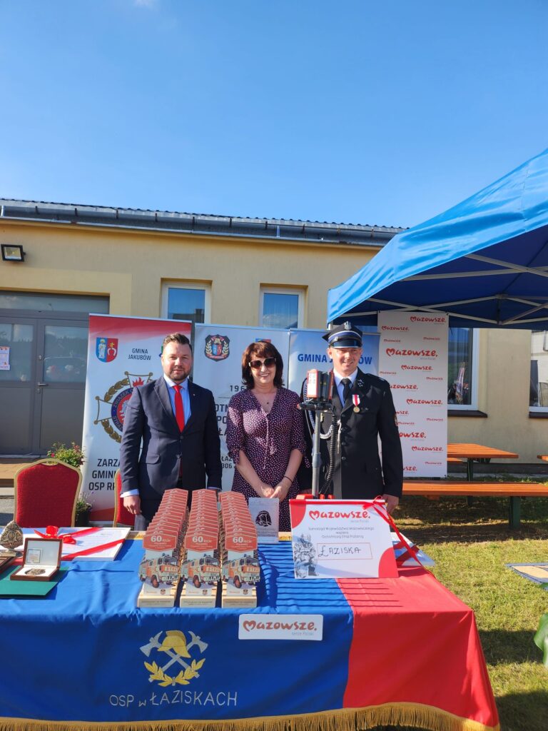 Na zdjęciu troje ludzi stoi przed stołem pokrytym niebiesko-czerwonym obrusem z logo OSP. W tle znajduje się jasnożółty budynek oraz niebieski namiot. Na stole widoczne są trofea i baner Mazowsze.
