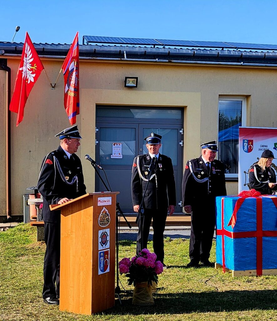 Trzech mężczyzn w mundurach strażackich stoi na trawie przed budynkiem. Obok znajduje się podium oraz duży niebieski pakunek przewiązany czerwoną wstążką. W tle widać flagi państwowe na tle jasnej ściany.