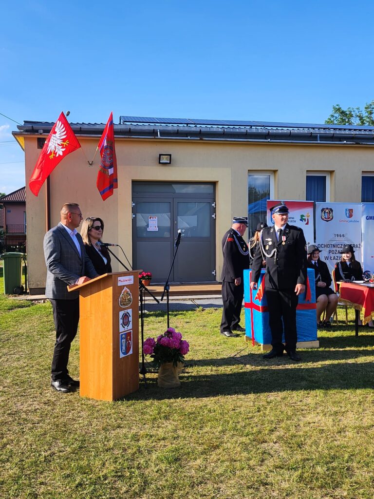 Na trawniku przed budynkiem stoi podium z mówcami. Tło zdobią czerwone flagi i banery. Obok znajdują się druhowie strażacy w mundurach.