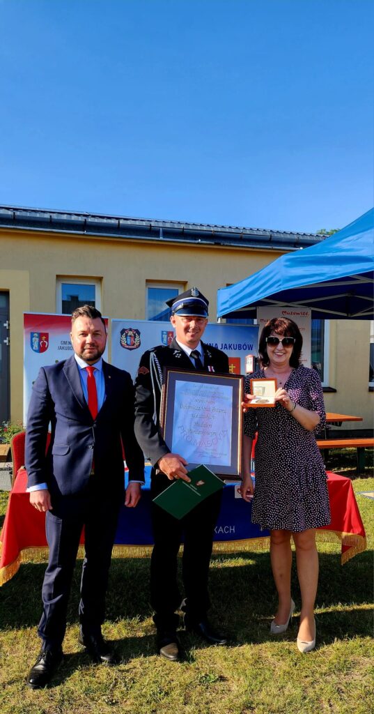 Trzy osoby stoją przed budynkiem na trawie. Dwie z nich trzymają certyfikaty i nagrodę. W tle niebieskie niebo i namiot.