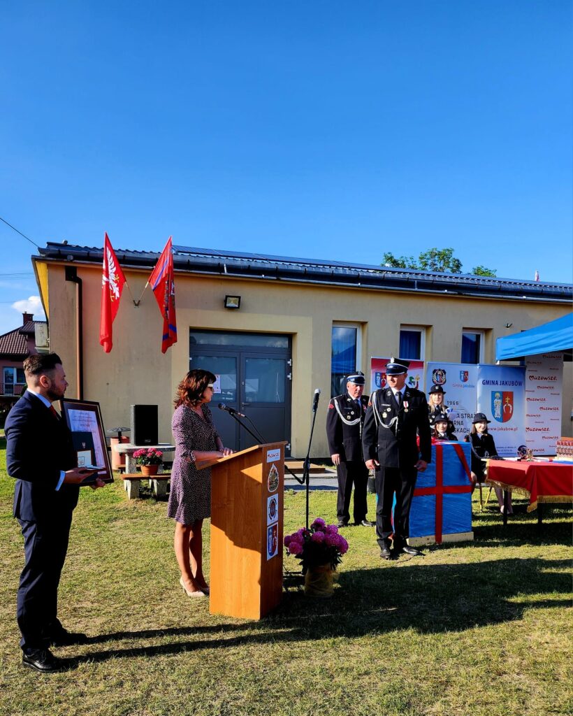 Na zdjęciu widać ceremonię przed małym budynkiem z czerwonymi flagami, na tle nieba. Kobieta przemawia przy drewnianym podium; obok niej stoją osoby w mundurach. W tle namiot z logo gminy i stoły z obrusami.