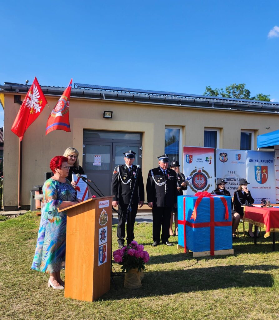 Na trawie, przed budynkiem z flagami, kobieta przemawia przy mównicy. Obok stoją strażacy w mundurach i duży prezent z kokardą. Tło jest jasne, z niebieskim niebem.