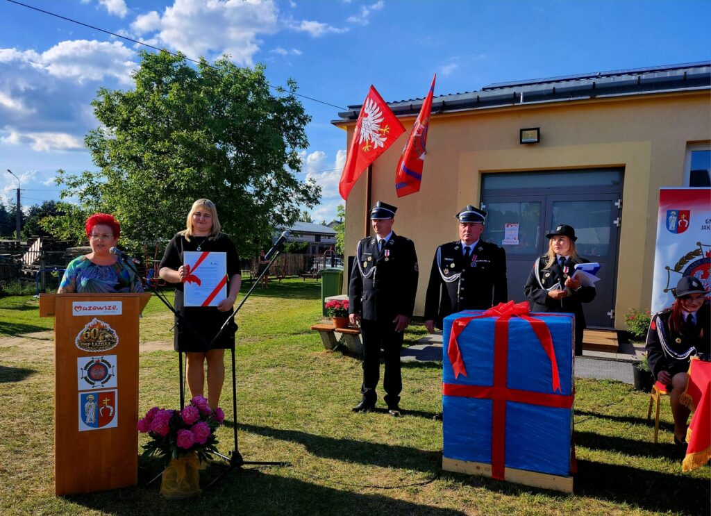Na zdjęciu grupa osób w eleganckich mundurach strażackich stoi przed budynkiem. Kobieta przemawia przy podium z mikrofonem. W tle jasne niebo, zielone drzewa i dwa czerwone sztandary.
