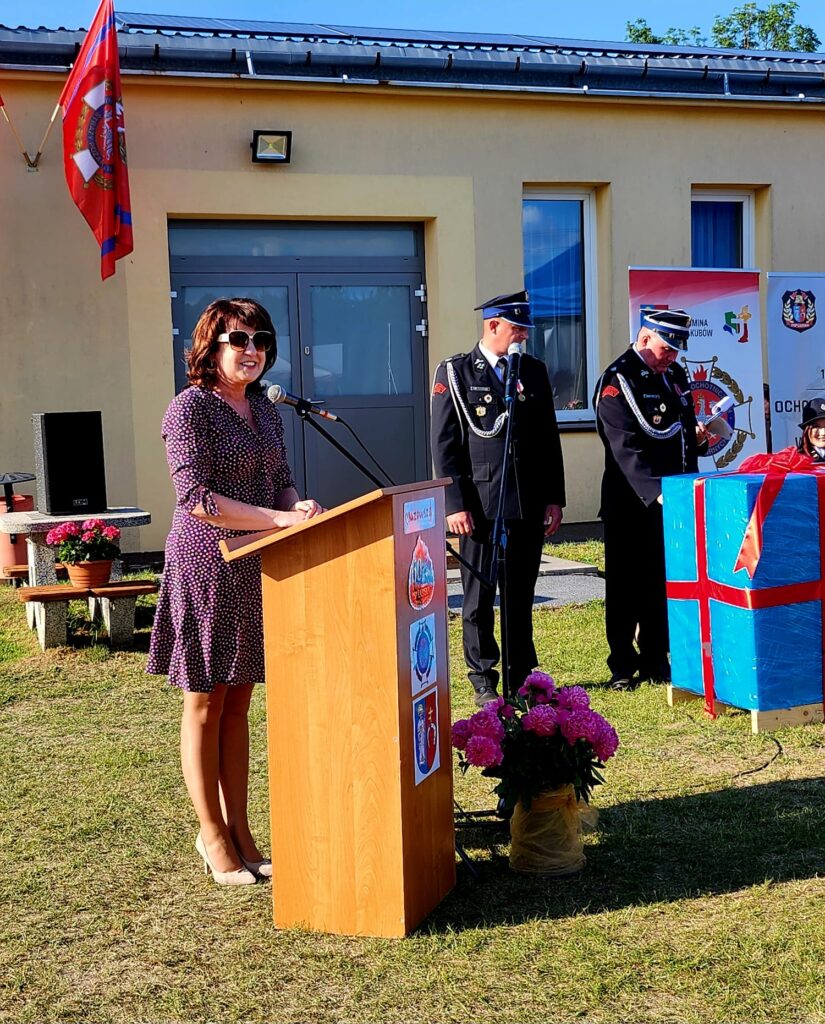 Kobieta w okularach przemawia przy drewnianej mównicy na tle żółtego budynku. Obok stoją dwaj strażacy w mundurach. Na trawie kwiaty i niebieskie pudełko z czerwoną wstążką.