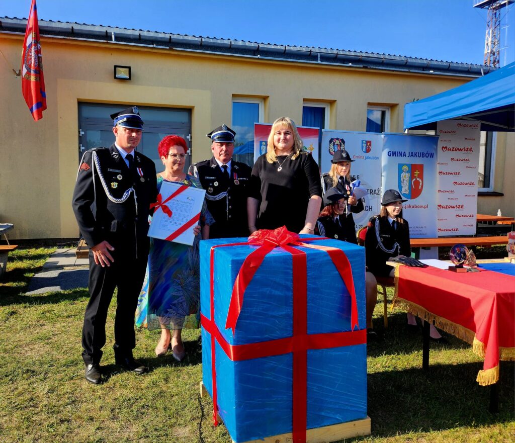 Grupa osób w mundurach strażackich stoi przy dużym niebieskim prezencie z czerwonymi wstążkami. Tło stanowi budynek z żółtą elewacją i flagami. Na stole obok znajdują się dekoracje, a wszystko odbywa się na zewnątrz przy