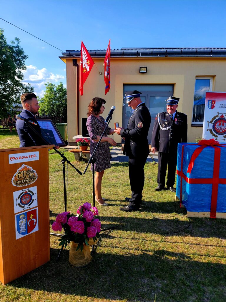 Na zdjęciu odbywa się ceremonia przed budynkiem z czerwonymi flagami. Kobieta gratuluje strażakowi w mundurze. Obok stoi mikrofon i bukiet różowych kwiatów.