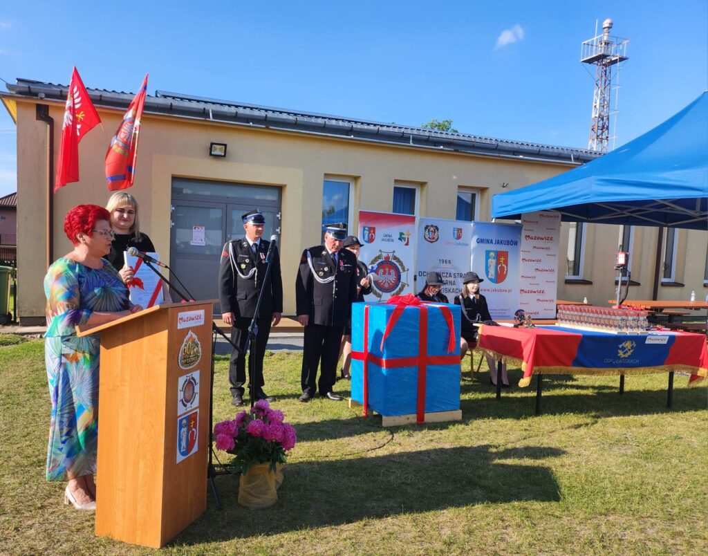 Na zdjęciu uroczystość na zewnątrz remizy strażackiej z żółtym budynkiem w tle. Kobieta przy mównicy, obok strażacy w mundurach i duży prezent owinięty w niebieski papier z czerwoną wstążką. Nieb