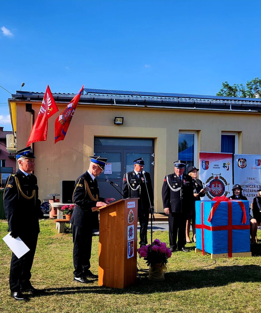 Na zdjęciu jest ceremonia strażaków w mundurach na tle budynku. Przód dekoruje podium i czerwone flagi, obok duże, zapakowane pudełko. Tło ma niebieskie niebo i drzwi budynku.