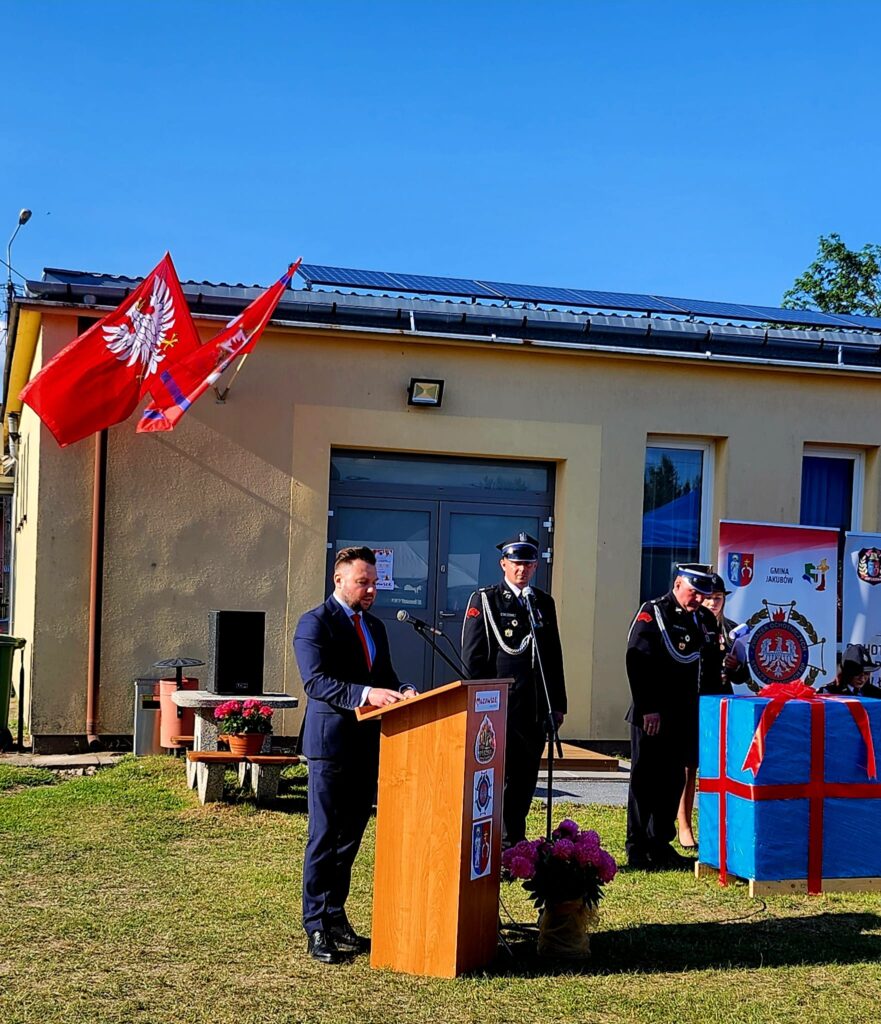 Na zdjęciu mężczyzna przemawia przy drewnianym podium, obok stoją strażacy. W tle budynek z flagami narodowymi i dużym, zapakowanym prezentem. Scena na zewnątrz, w słoneczny dzień, na trawie.