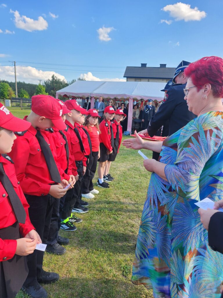 Grupa dzieci w czerwonych mundurach stoi na trawie. Obok osoby w formalnym stroju. Tło: niebo, namiot, budynek.