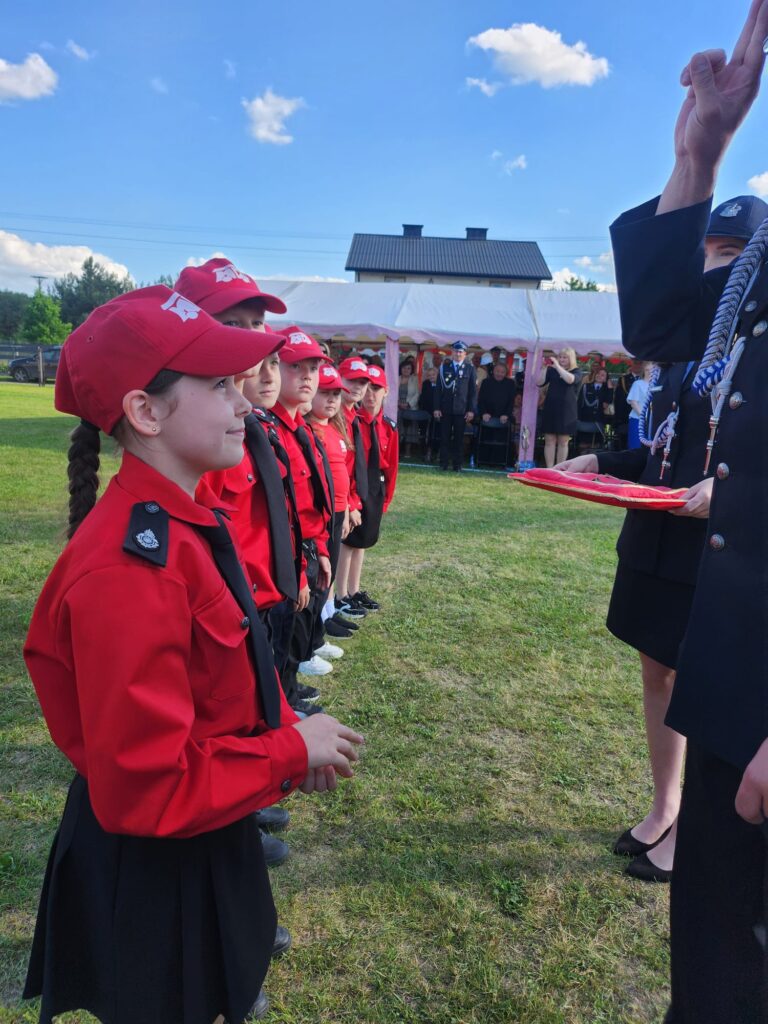 Na zdjęciu grupa dzieci w czerwonych mundurach stoi w szeregu na zielonej trawie. W tle biały namiot i niebieskie niebo z chmurami. Osoba z lewej ręką uniesioną do przysięgi.