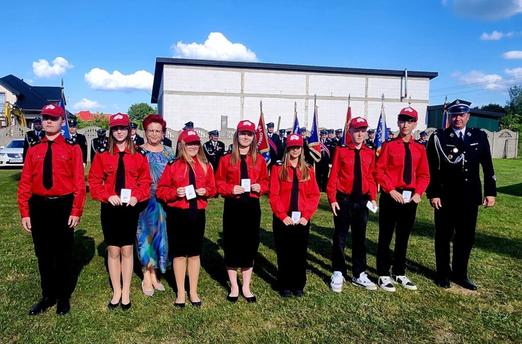 Grupa młodych ludzi w czerwonych koszulach i czapkach stoi na trawie. Za nimi znajduje się formacja w galowych mundurach z flagami. Tło stanowi budynek i niebieskie niebo z białymi chmurami.