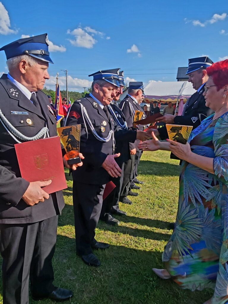 Czterech strażaków w galowych mundurach odbiera nagrody na trawiastym tle. Kobieta w kolorowej sukni wręcza im dyplomy. Jasne, słoneczne niebo z chmurami w tle.