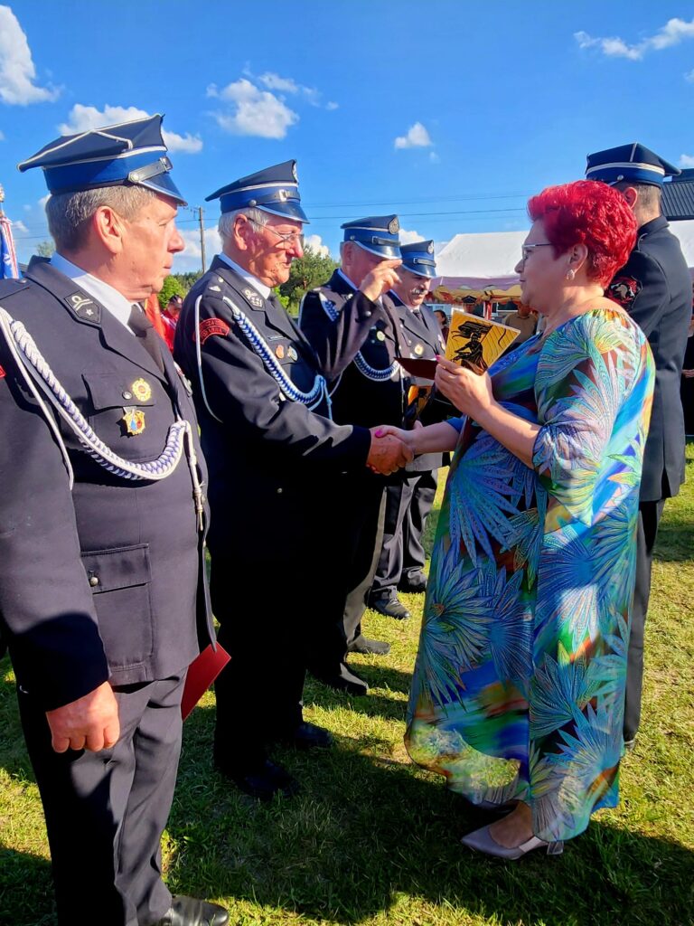 Grupa strażaków w mundurach odbiera nagrodę od kobiety w kolorowej sukni. Tło to zielona trawa, niebieskie niebo z chmurami. Świeci słońce, tworząc jasną atmosferę.