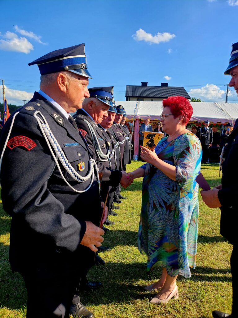 Na zdjęciu widać grupę strażaków w ciemnych mundurach i kobietę w kolorowej sukience, która wręcza nagrodę. W tle jest namiot i dom, a nad nimi błękitne niebo z białymi chmurami. Scena odbywa się na trawie
