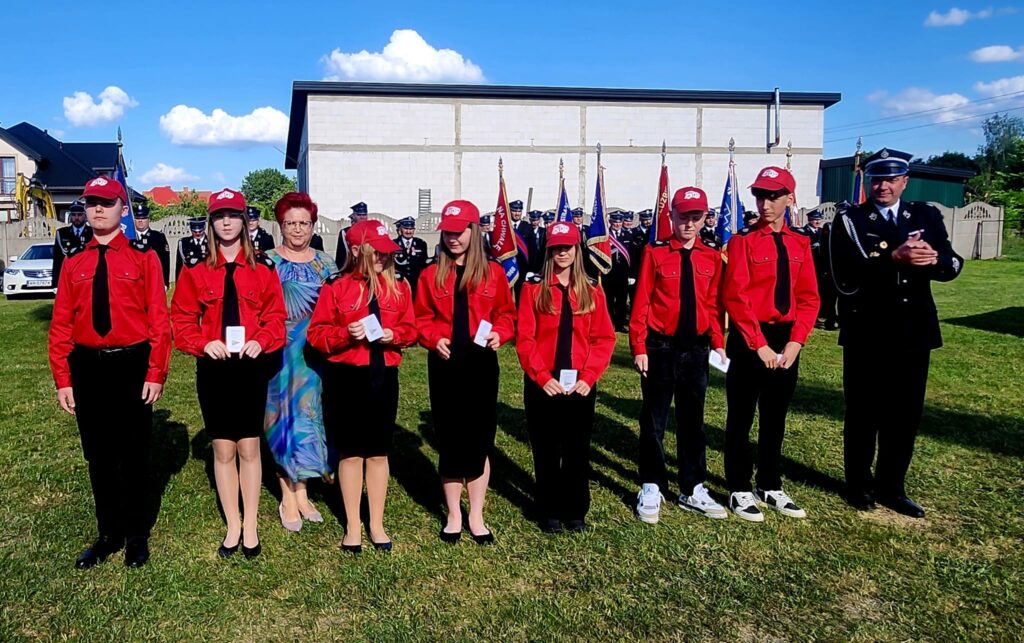 Grupa młodych osób w czerwonych uniformach i czapkach stoi na trawie. Za nimi widoczni strażacy w mundurach z flagami. W tle jasny budynek i niebieskie niebo z chmurami.