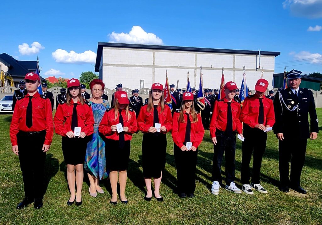 Na zdjęciu grupa młodzieży w czerwonych koszulach i czapeczkach stojąca na trawie. W tle widać budynek i osoby w mundurach oraz flagi. Jasne niebo z puszystymi chmurami.