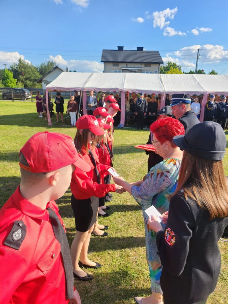 Grupa młodzieży w czerwonych uniformach otrzymuje nagrody na zielonej trawie. W tle biały namiot oraz zebrani goście. Niebo błękitne z chmurkami.