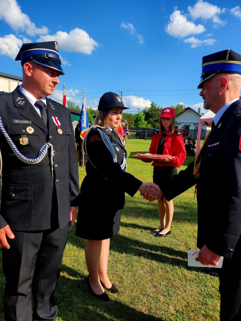 Grupa strażaków w mundurach na zielonej trawie podczas ceremonii. Niebieskie niebo z chmurami w tle. Kobieta w czerwonym stroju trzyma tacę.