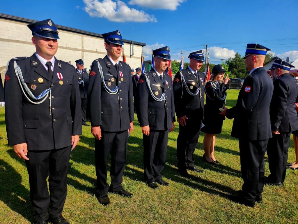 Grupa strażaków w mundurach stojących na zielonej trawie. Niebieskie niebo i biały budynek w tle. Ceremonia z odznaczeniami.