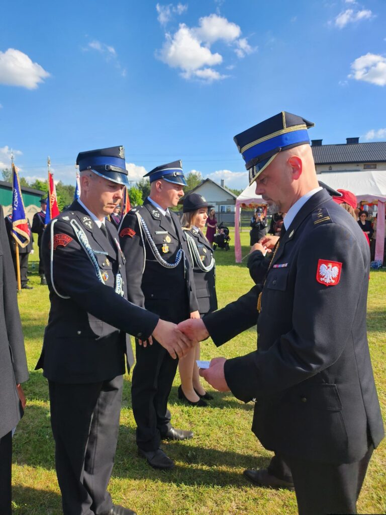 Na zdjęciu widoczni są strażacy w czarnych mundurach, wymieniający uścisk dłoni. Tło to niebieskie niebo z chmurami i zielona trawa. W tle widać namiot i budynek.