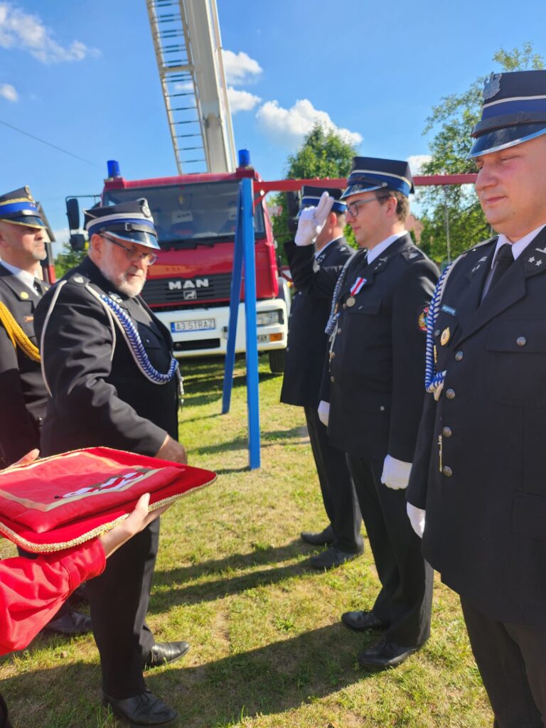 Na zdjęciu strażacy w mundurach salutują przed czerwonym wozem strażackim. Trzymają sztandar na czerwonej poduszce. Tło stanowi jasne, słoneczne niebo i drzewa.