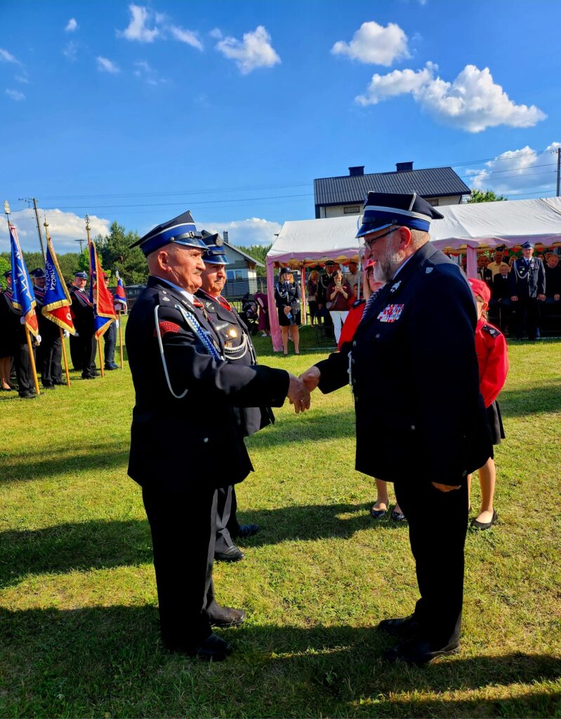 Dwaj mężczyźni w mundurach strażackich podają sobie ręce na trawniku. W tle namiot i ludzie, niebo jest błękitne z chmurami. Widoczne są też sztandary i budynek.