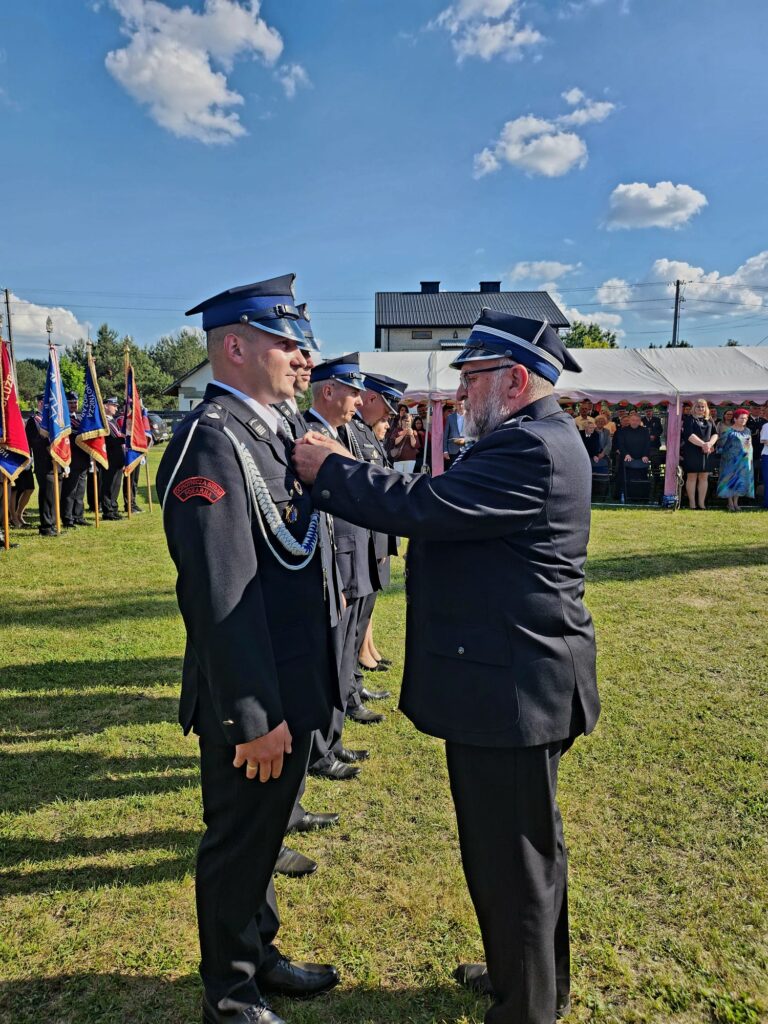 Ceremonia odznaczania strażaków na zielonym trawniku. Mężczyźni w ciemnych mundurach, niebo z białymi chmurami w tle. W tle namiot i publiczność.