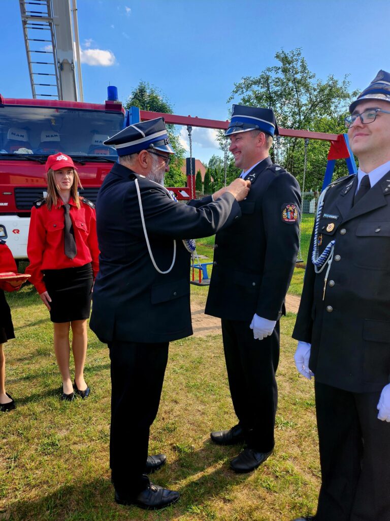 Na zdjęciu widać strażaków w mundurach podczas ceremonii. W tle widać czerwony wóz strażacki i błękitne niebo. Jeden strażak przypina medal drugiemu.