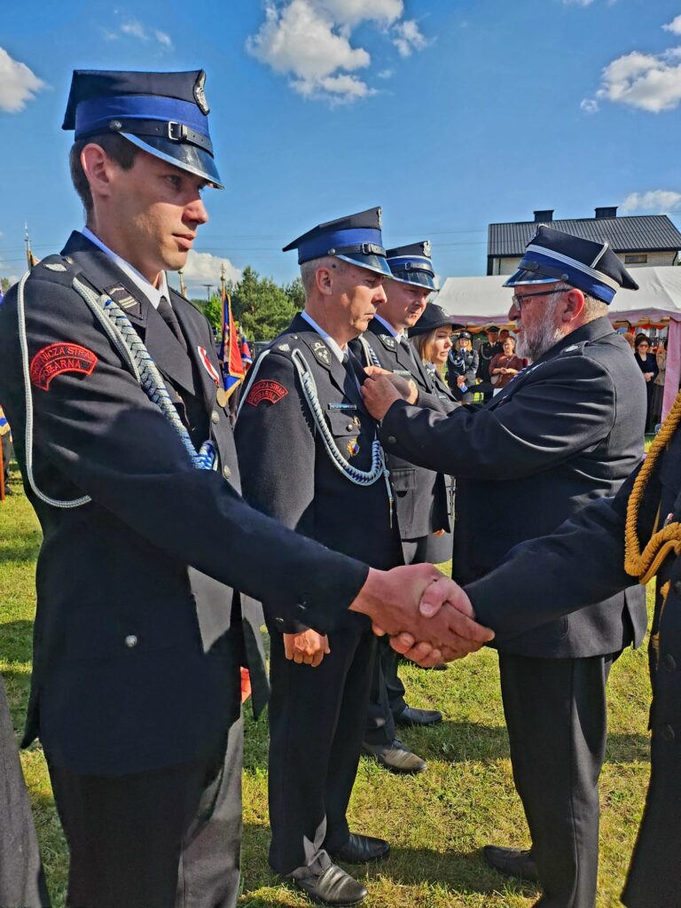Na zdjęciu grupa strażaków w mundurach, błękitne niebo z chmurami w tle. Jeden strażak odznacza drugiego, wymieniając uścisk dłoni. Za nimi widać namiot i budynek.