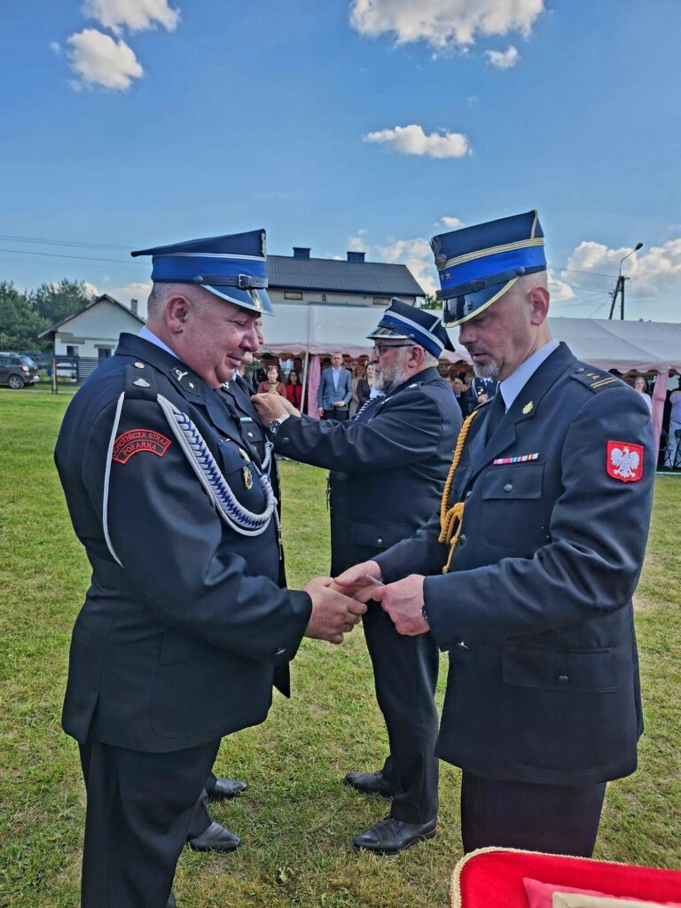 Dwóch strażaków w granatowych mundurach ściska się, drugi medal wręcza; niebo niebieskie z chmurkami, namioty w tle.