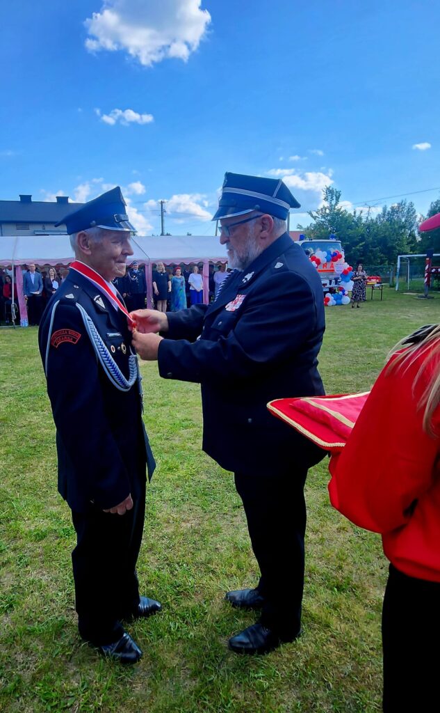 Dwóch strażaków w mundurach, jeden przypina medal drugiemu na zielonej trawie. Niebieskie niebo z chmurami. W tle namioty i ludzie.