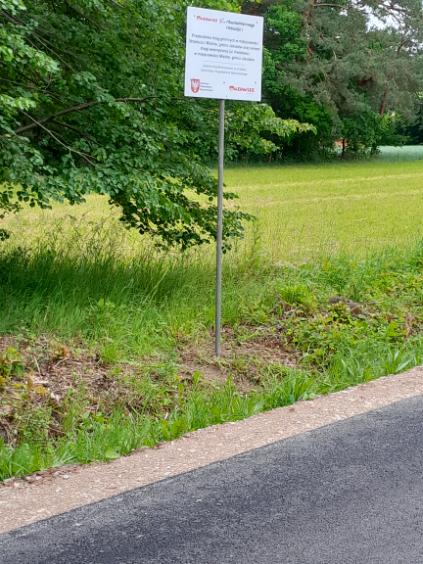 Znajduje się tu znak na metalowym słupku przy asfaltowej drodze. W tle zielone drzewa i trawiaste pole. Kolory to zielony, szary i brązowy.