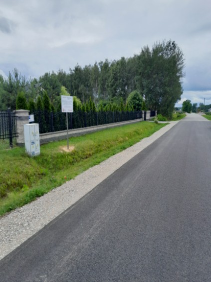 Droga asfaltowa wiedzie obok zielonego żywopłotu i metalowego ogrodzenia. Obok stoi tablica informacyjna i szary słupek. Tło zdobi zadrzewiony krajobraz pod pochmurnym niebem.