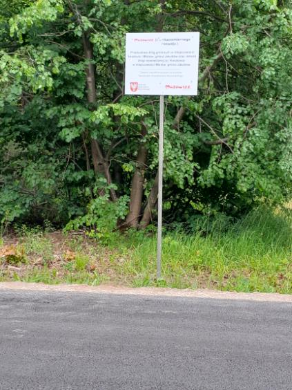 Znak na poboczu drogi, na tle zielonych liści i krzewów, zawiera tekst i logo z czerwonym akcentem, stojący na metalowym słupku.