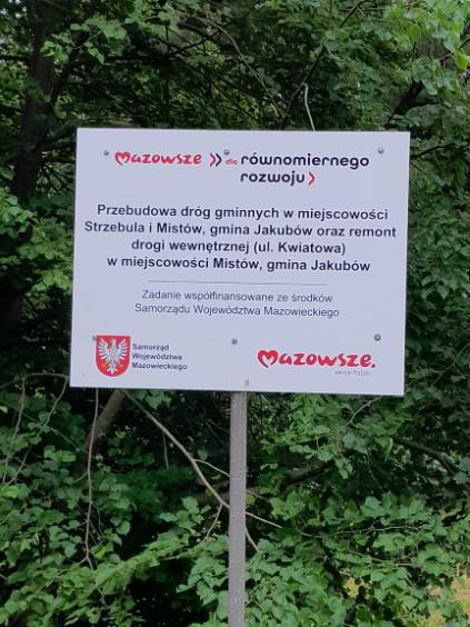 Tablica informacyjna na tle zielonych drzew. Białe tło z czerwonym logo Mazowsze i czarnym tekstem o przebudowie dróg. Projekt współfinansowany przez Samorząd Województwa Mazowieckiego.