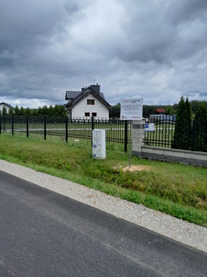 Dom w bieli z ciemnym dachem za czarnym płotem. Przed płotem trawa, znaki i szosa. Nad domem szare, pochmurne niebo.
