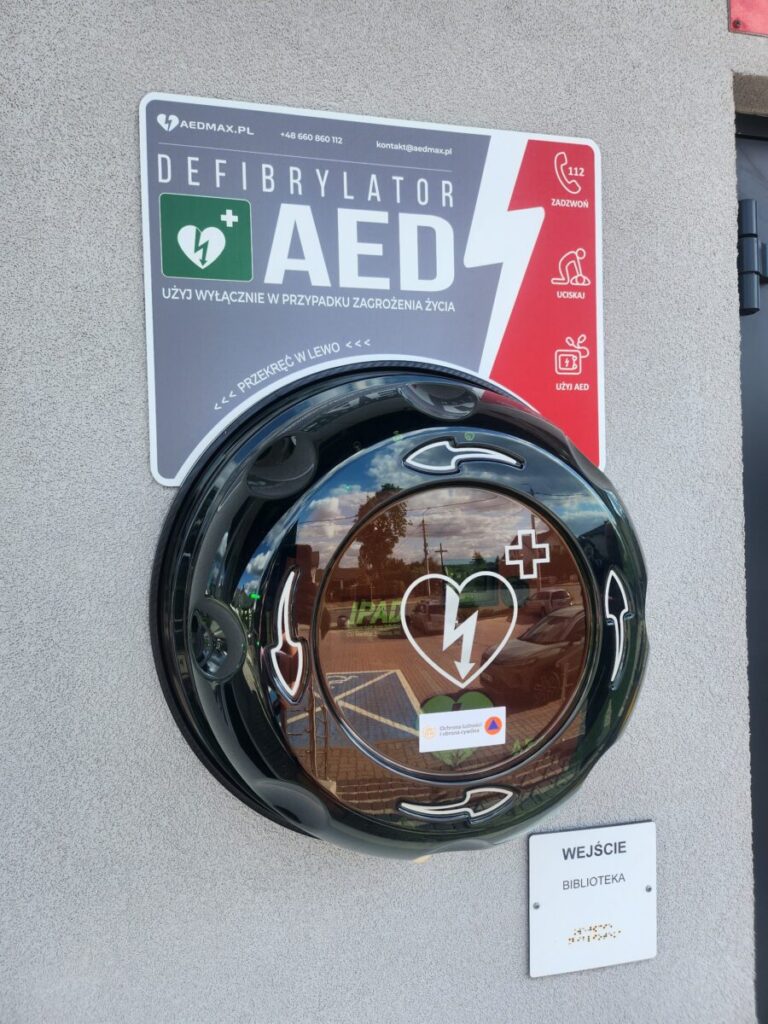 Zdjęcie przedstawia defibrylator AED w szarej obudowie na tle ściany. Widoczna jest tablica informacyjna z czerwonym akcentem. Obok znajduje się znak wejścia do biblioteki.